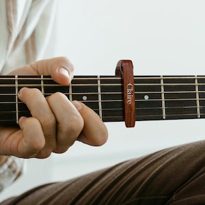 May include: A wooden guitar capo with the name "Claire" engraved on it. The capo is attached to the fretboard of an acoustic guitar.