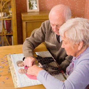 May include: An elderly couple working together to complete a jigsaw puzzle at a wooden table. The puzzle features a photograph of two people. A framed wedding photo is visible in the background. The couple is focused on the puzzle.