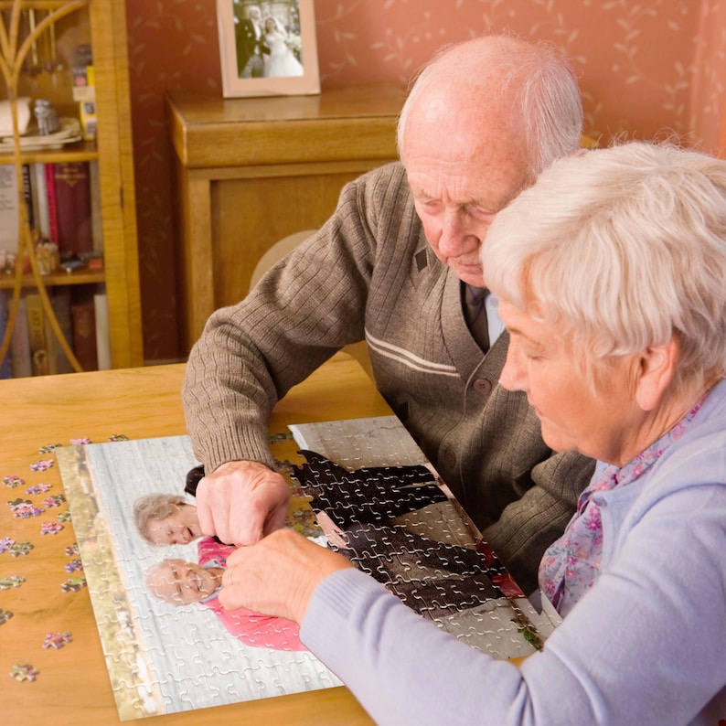 May include: An elderly couple working together to complete a jigsaw puzzle at a wooden table. The puzzle features a photograph of two people. A framed wedding photo is visible in the background. The couple is focused on the puzzle.