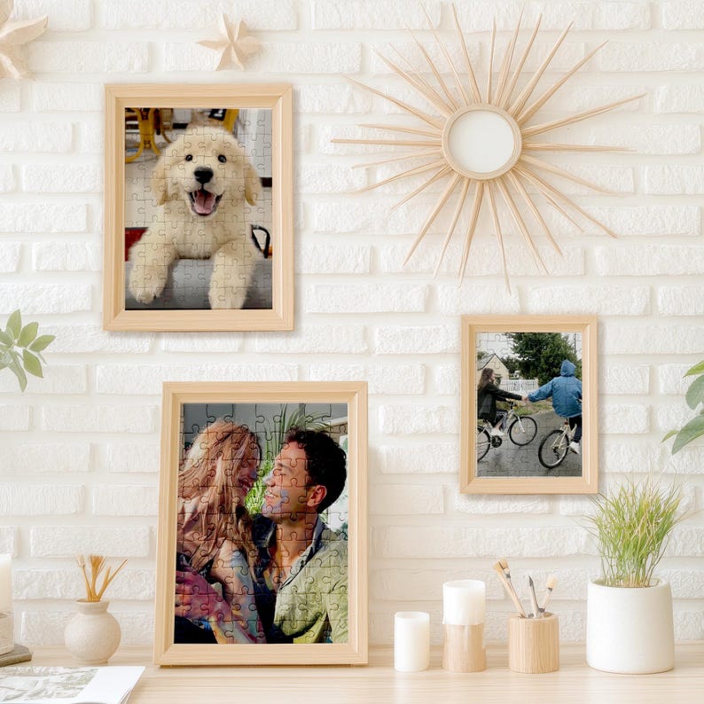 May include: A collection of framed jigsaw puzzles with light wood frames. One puzzle features a smiling golden retriever, another a couple, and a third shows two people on bicycles. The puzzles are displayed on a white brick wall.