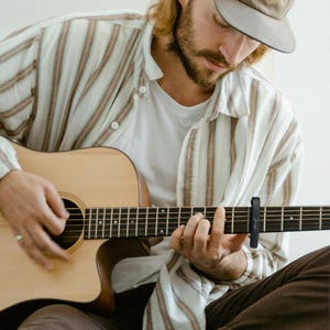 May include: A person playing an acoustic guitar with a capo on the second fret. The guitar is brown with a natural finish. The person is wearing a striped shirt and a baseball cap.