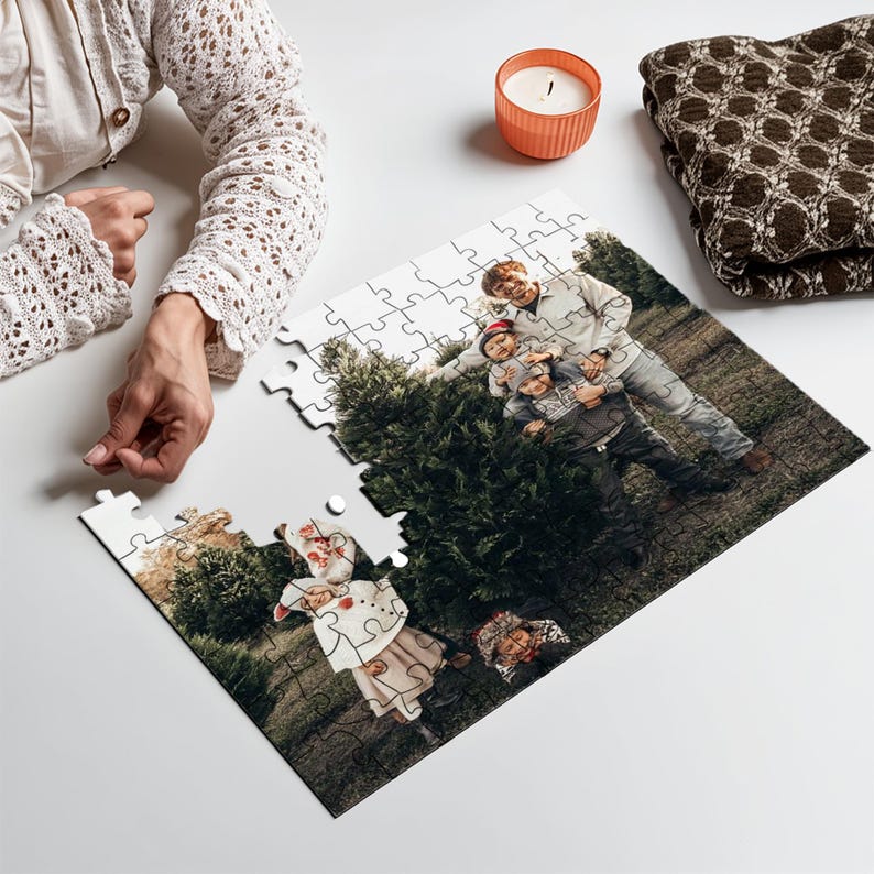 May include: A partially assembled jigsaw puzzle featuring a family portrait in a forest setting. The puzzle pieces are white with a glossy finish. A lit orange candle and a folded brown blanket are also visible.