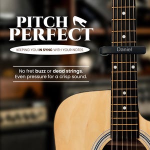 May include: A black guitar capo with the word "Daniel" engraved on it. The capo is attached to the neck of an acoustic guitar. The text "Pitch Perfect" is displayed above the guitar, along with the tagline "Keeping you in sync with your notes".