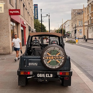 Peut inclure: Un v&eacute;hicule Suzuki gris fonc&eacute; avec une capote marron, dot&eacute; d'une housse de roue de secours avec un graphique de globe et le texte STUSSY, PEACE & PROSPERITY NEGRIL JAMAICA, et STUSSY BEACH BAR N SURF SHACK SINCE 1980. La plaque d'immatriculation est G615 ELB.