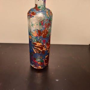 May include: A decorative glass bottle with a colorful, abstract design. The bottle features a blue base with swirls of red, orange, green, and gold paint. The neck of the bottle is clear with blue accents.