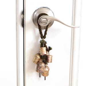 May include: A cluster of small, weathered brass-colored metal bells suspended from a dark brown braided cord. The bells feature a rustic, aged patina with visible metallic textures and are attached to a silver-toned metal door handle.