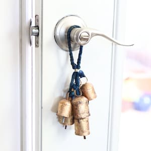 May include: A cluster of small, aged brass bells hangs from a dark blue braided cord, attached to a silver door handle. The bells have a rustic, weathered appearance, and the cord is knotted at the top.