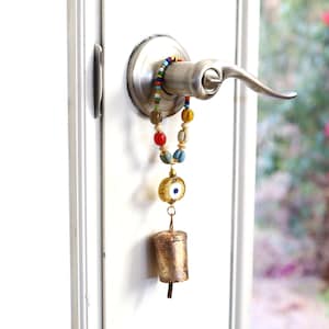 May include: A decorative door handle charm with a beaded strand in various colors, including red, blue, and yellow. The charm features a gold-colored eye pendant and a small, aged bell. The charm is attached to a silver-colored door handle.
