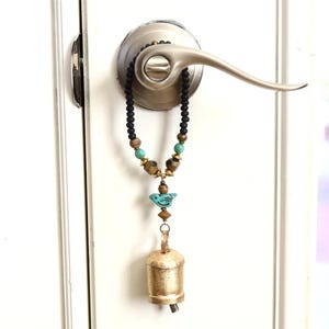 May include: A decorative door hanger featuring a beaded necklace with turquoise and gold accents, a small blue bird charm, and a gold-colored bell. The door hanger is attached to a silver door handle and hangs against a white door.
