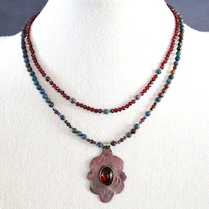 May include: A two-strand necklace with a floral pendant. The top strand is composed of small red beads, while the bottom strand features a mix of blue, green, and brown beads. The pendant is a silver-toned metal with a red oval stone.