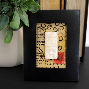 May include: A small black rectangular frame containing a cream-colored carved face relief centered on a background of tan paper with black circular ink stamps and a small red postage stamp with the word India printed in the bottom right corner.