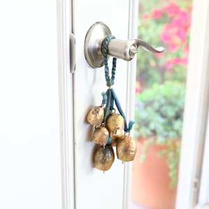 May include: A silver door handle with a decorative string of gold bells hanging from it. The bells are tied together with blue and green string.