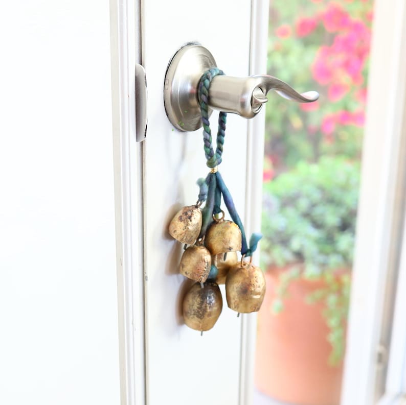May include: A silver door handle with a decorative string of gold bells hanging from it. The bells are tied together with blue and green string.