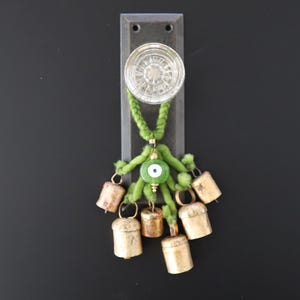 May include: A clear glass doorknob is mounted on a dark metal plate. A green braided cord with a green eye charm and small brass bells hangs from the doorknob. The bells are various sizes and shapes.