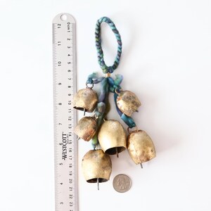 May include: A set of seven gold-colored bells hanging from a blue and green braided cord. The bells are various sizes and have a worn, antique look. A ruler is in the background for scale.