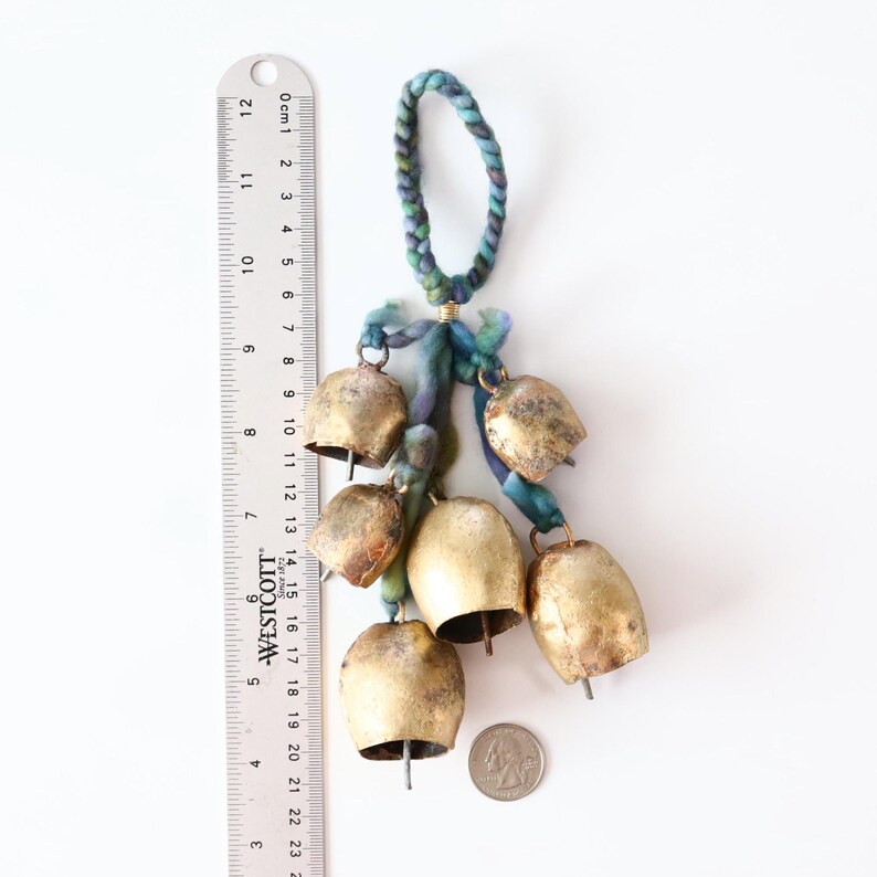 May include: A set of seven gold-colored bells hanging from a blue and green braided cord. The bells are various sizes and have a worn, antique look. A ruler is in the background for scale.