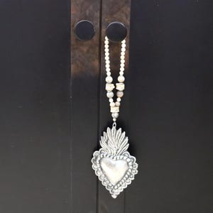 May include: A silver-toned pendant necklace featuring a heart-shaped design with a flame motif. The pendant hangs from a beaded strand of white and tan beads. The necklace is displayed against a dark background.
