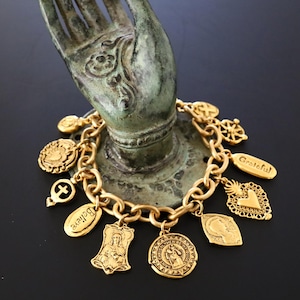 May include: Gold-toned charm bracelet featuring various religious and inspirational charms. Charms include crosses, medallions, and the word "Grateful." The bracelet is displayed on a bronze hand sculpture against a black background.