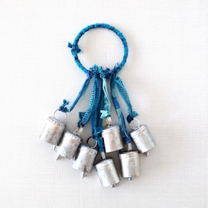 May include: A cluster of seven silver-colored bells suspended from a blue fabric loop and ribbons. The bells are cylindrical with a small clapper. The loop and ribbons are various shades of blue, creating a decorative hanging ornament.