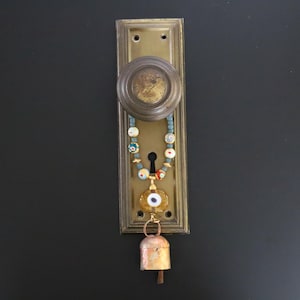May include: A vintage brass door plate and round knob featuring a decorative hanging ornament. The ornament consists of a string of multi-colored glass beads, a central yellow glass evil eye charm, and a small weathered metal bell at the bottom.