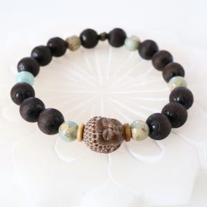 May include: A brown wooden beaded bracelet with a brown Buddha head charm and light blue and beige beads.