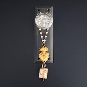 May include: A rectangular dark metal wall plate featuring a clear glass knob at the top. A string of small white beads hangs from the knob, connecting to a gold-toned heart-shaped pendant with a cross and a small metallic bell at the bottom.