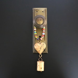 May include: A brass door knob and rectangular backplate featuring a decorative hanging ornament. The ornament consists of a string of multicolored glass beads attached to a textured metal heart charm and a small cylindrical brass bell.