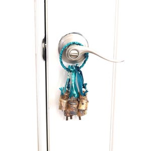 May include: A cluster of small, aged brass bells with a turquoise and blue fabric loop and ribbons, hanging from a silver door handle. The bells have a rustic, weathered appearance, and the fabric adds a pop of color.