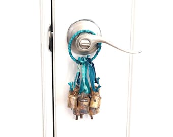 May include: A cluster of small, aged brass bells with a turquoise and blue fabric loop and ribbons, hanging from a silver door handle. The bells have a rustic, weathered appearance, and the fabric adds a pop of color.