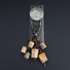 May include: A clear glass door knob mounted on a rectangular dark metal backplate. A braided black and white patterned fabric strap hangs from the knob, featuring five weathered, rustic brass-toned metal bells attached at varying lengths.