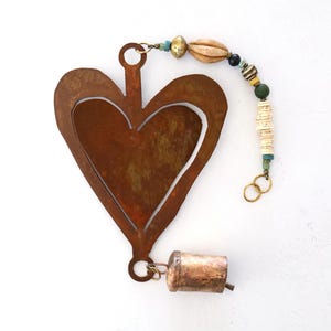 May include: A rustic, heart-shaped metal ornament with a cutout heart design. It features a small bell and a beaded strand with various colors and textures. The ornament is approximately 6 inches in length.