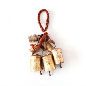 May include: A set of five small, gold-colored bells hanging from a red and brown braided cord. The bells are tied together with the cord and have a rustic, vintage look.