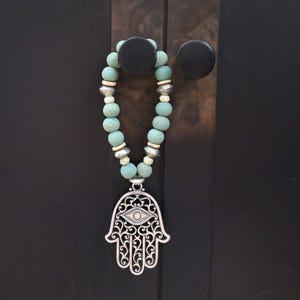 May include: A turquoise beaded necklace with a silver Hamsa hand pendant. The pendant features an eye design and intricate openwork. The necklace is hanging on a dark brown surface with two black knobs.