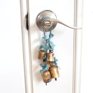 May include: A cluster of four weathered, cylindrical brass bells suspended by frayed, light blue fabric ribbons. The bells feature a tarnished, metallic finish and are tied together in a decorative arrangement, hanging from a silver-toned door handle.