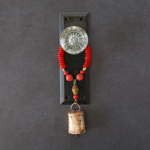 May include: A vintage-style door knocker with a clear glass knob and a dark metal backplate. A red beaded strand with gold accents hangs from the knob, leading to a small, aged brass bell. The background is a dark gray.