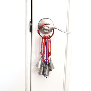 May include: A decorative door knocker with a ring of colorful fabric and hanging silver bells. The fabric includes shades of red, orange, and purple. The bells are attached to the fabric ring and hang down.