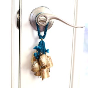May include: A cluster of small, gold-colored bells hangs from a teal blue braided cord, attached to a silver door handle. The bells have a rustic, textured appearance, suggesting a handmade craft item.