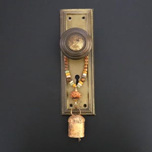 May include: A vintage brass door plate with a round knob and a keyhole. A decorative hanging ornament is attached to the knob, featuring a string of multi-colored beads, a carved peach-colored floral bead, and a small rustic metal bell at the end.