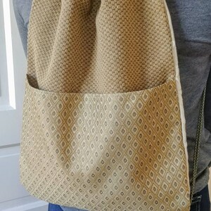 May include: A tan and gold patterned drawstring backpack with a front pocket. The backpack has a diamond pattern and green rope straps. The bag is worn over a gray shirt and blue jeans.