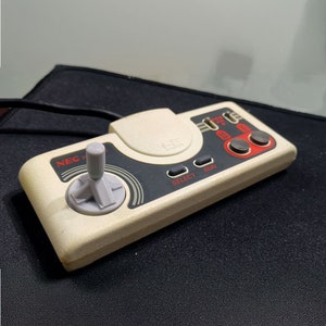 Könnte beinhalten: Ein weißer und grauer NEC PC-Engine-Controller mit einem Joystick, vier Knöpfen und den Worten "SELECT" und "RUN" auf der Vorderseite.