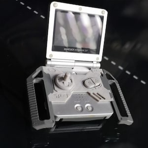 May include: Silver Game Boy Advance SP handheld game console with a flip-up screen. The console features a directional pad, A and B buttons, and start/select buttons. The screen displays the text "GAME BOY ADVANCE SP".