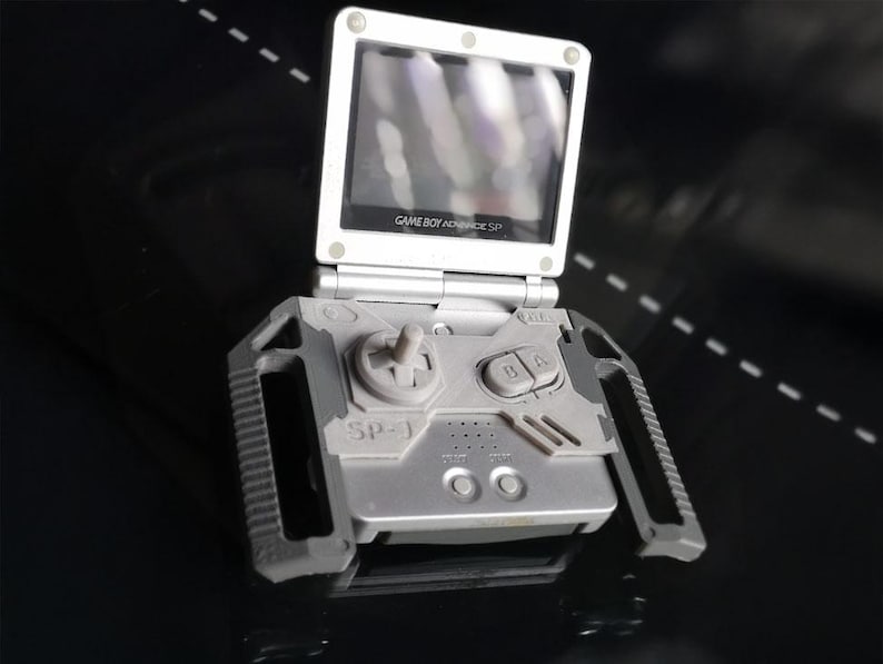 May include: Silver Game Boy Advance SP handheld game console with a flip-up screen. The console features a directional pad, A and B buttons, and start/select buttons. The screen displays the text "GAME BOY ADVANCE SP".