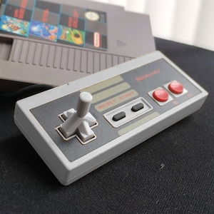 May include: A grey Nintendo Entertainment System controller with a white joystick and two red buttons labelled "Start" and "Select". The controller is on a black surface.