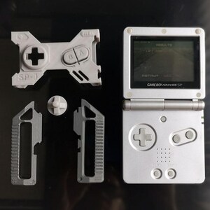 May include: A silver Game Boy Advance SP handheld console with a screen displaying a game. Gray replacement parts, including a directional pad, buttons, and side grips, are arranged around it. The text "SP-1" is visible on one of the parts.