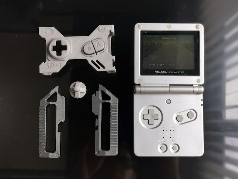 May include: A silver Game Boy Advance SP handheld console with a screen displaying a game. Gray replacement parts, including a directional pad, buttons, and side grips, are arranged around it. The text "SP-1" is visible on one of the parts.