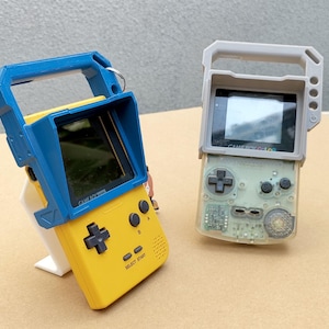 Könnte beinhalten: Zwei tragbare Videospielkonsolen mit kundenspezifischen 3D-gedruckten Gehäusen. Die Konsole links ist gelb mit einem blauen Sonnenschutz und dem Text "GAME BOY COLOR" auf der Vorderseite. Die Konsole rechts ist grau mit einem klaren Bildschirm und dem Text "GAME BOY COLOR" auf der Vorderseite.