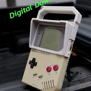 May include: A white and beige Game Boy handheld video game console with a white protective case. The case has a handle and a visor to block out light. The Game Boy has a black directional pad, two pink buttons, and the text "Nintendo GAME BOY" printed on the front.