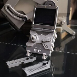 May include: A gray Game Boy Advance SP-0 handheld gaming console with a flip-up screen. The console has a directional pad, A and B buttons, and a rectangular design. Two gray accessories are in front of the console.