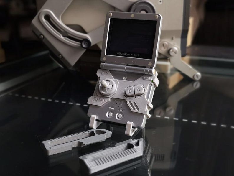 May include: A gray Game Boy Advance SP-0 handheld gaming console with a flip-up screen. The console has a directional pad, A and B buttons, and a rectangular design. Two gray accessories are in front of the console.