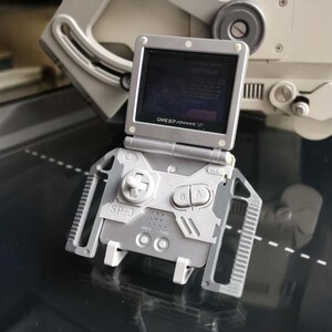 May include: A silver Game Boy Advance SP handheld console with a custom SP-J case. The screen is open, displaying a game. The case has a D-pad, A and B buttons, and the text "SP-J". The console is on a reflective surface.